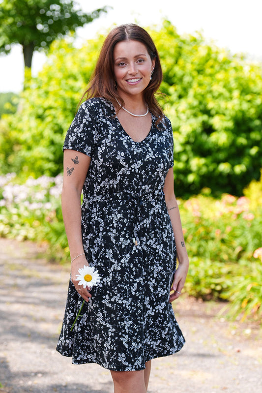 
A woman wearing the Mambo Dress from Rien ne se Perd, a black and white floral dress with a V-neck, short sleeves, adjustable empire waist, and knee-length A-line skirt. She is standing outside in a garden. 
