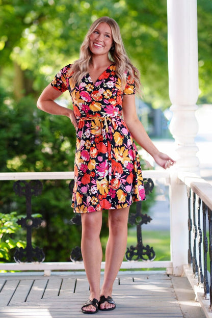 A woman wearing the Margarita Dress by Rien ne se Perd in Red/Orange floral print, with a faux wrap neckline, short sleeves, and short a-line skirt. She is standing on a porch. 