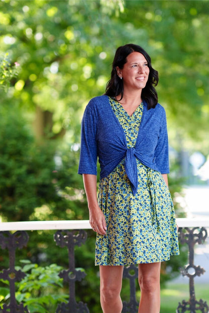 A woman wearing the Margarita Dress by Rien ne se Perd in Blue/Green floral print, with a faux wrap neckline, short sleeves, and short a-line skirt. She wearing it with a blue bolero and standing on a porch. 