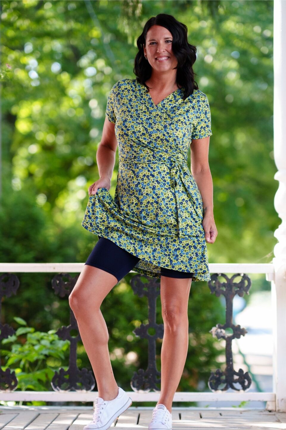 A woman wearing the Margarita Dress by Rien ne se Perd in Blue/Green floral print, with a faux wrap neckline, short sleeves, and short a-line skirt. She is wearing it with black bike shorts and standing on a porch. 