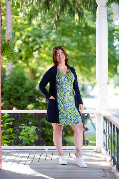 A woman wearing the Margarita Dress by Rien ne se Perd in Blue/Green floral print, with a faux wrap neckline, short sleeves, and short a-line skirt. She is wearing it with a navy cardigan and standing on a porch. 