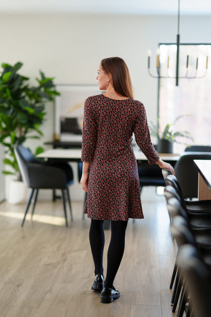 Back view of a woman wearing the Mattawin Dress from Rien ne se Perd in Red Floral, an above-the-knee length faux-wrap dress with three-quarter sleeves. She is standing in a modern, softly blurred dining room setting.
