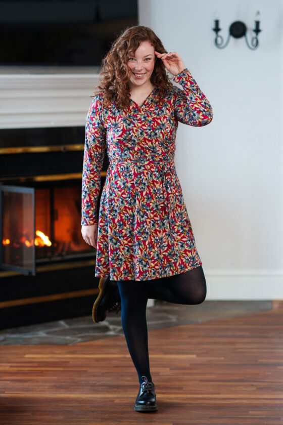 A woman wearing the Renoir Dress by Rien ne se Perd in Red Floral, standing in front of a fireplace