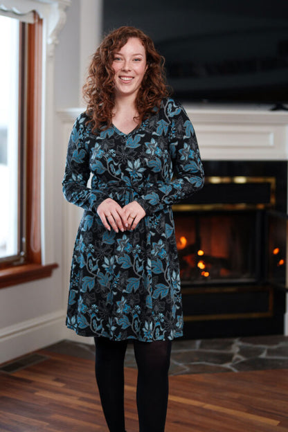 A woman wearing the Riopelle Dress by Rien ne se Perd in a Black/Turquoise print, standing in front of a fireplace