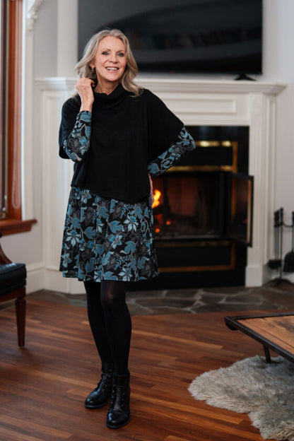 A woman wearing the Riopelle Dress by Rien ne se Perd in a Black/Turquoise print, witih the Delacroix Bolero-Poncho, standing in front of a fireplace