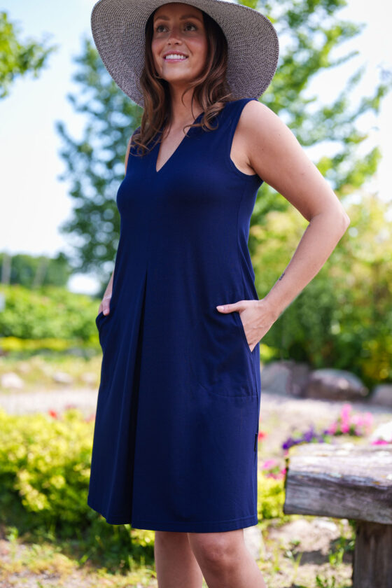 3/4 view of a woman wearing the Samba Dress by Rien ne se Perd in Navy, a sleeveless short dress with a V-neck, centre pleat, and side pockets. She is standing outside.