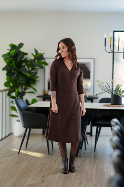 A woman wearing the St-Laurent Dress by Rien ne se Perd in Chocolate, a midi-length wrap style dress with a 3/4 sleeves and subtle shoulder pleats. She is standing in a dining room in front of a table. 