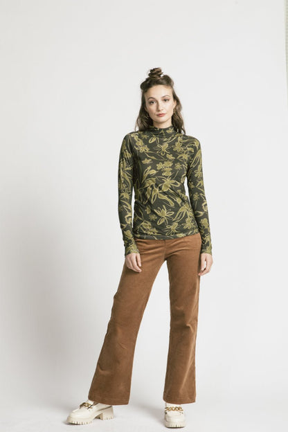 A woman wearing the Rebecca Turtleneck by Allison Wonderland in Green Floral with brown pants, standing in front of a white background 