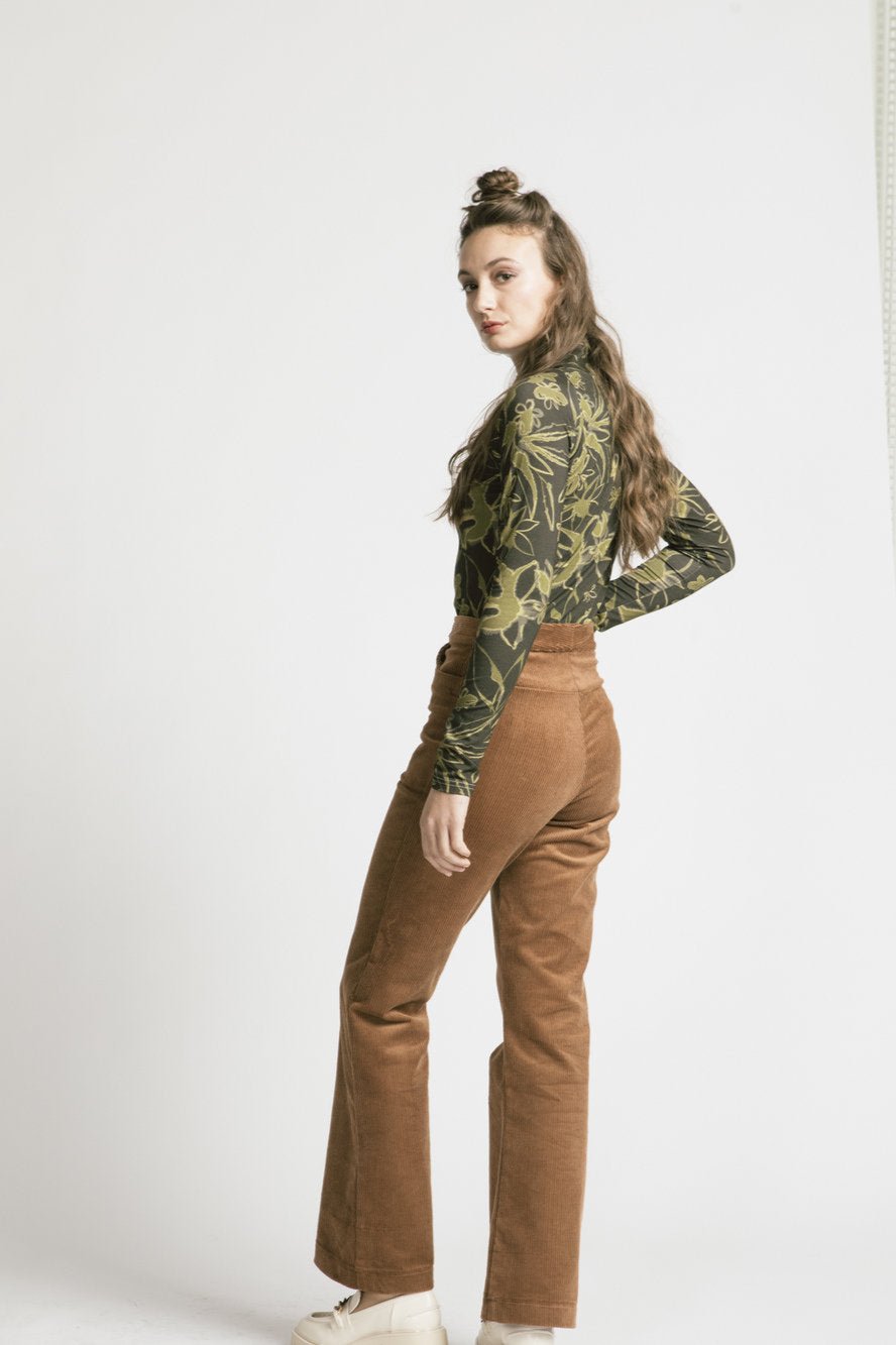 Back view of a woman wearing the Rebecca Turtleneck by Allison Wonderland in Green Floral with brown pants, standing in front of a white background 