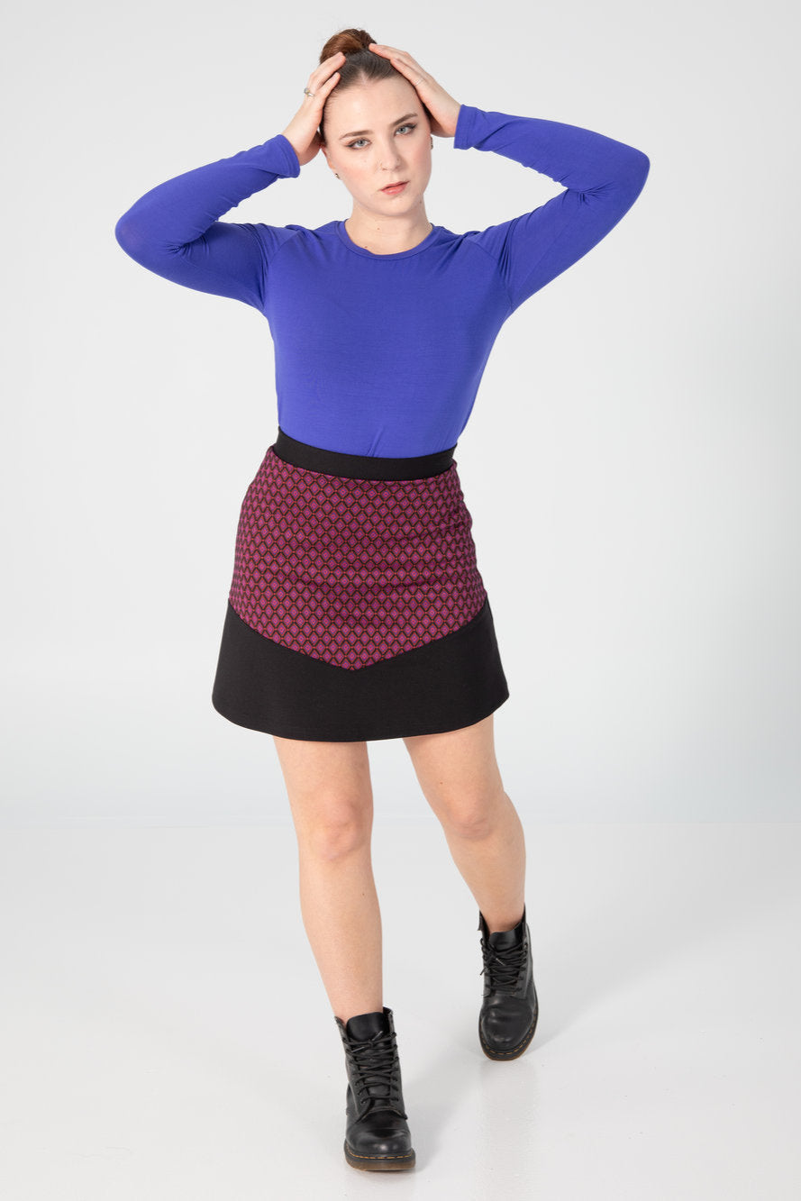 A woman wearing the A25 Sweater by Slak in Indigo, a long sleeved top with a round neck and a slim fit. She is wearing it with a pink and black skirt and standing in front of a white background. 