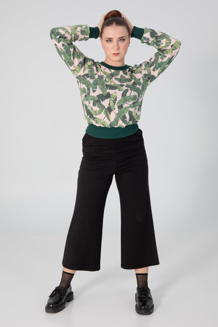A woman wearing the A25 Pants by Slak in Black, wide legged cropped pants with an elastic waist in Ponte Di Roma fabric. She is wearing them with  the Wanda Sweater by Slak in Pink and Green Tropical Leaves and standing in front of a grey background. 