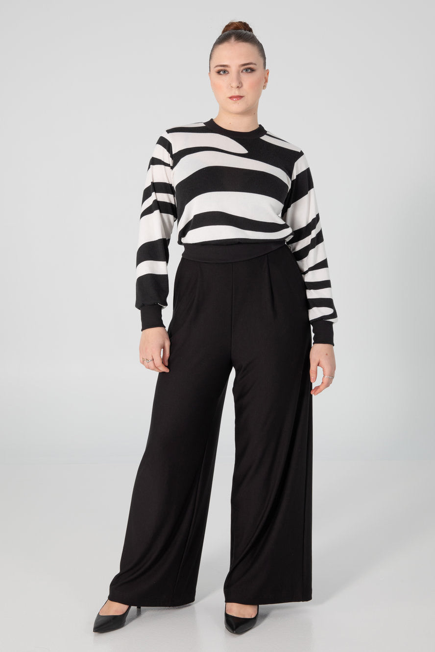 A woman wearing the Wanda Sweater by Slak in Black and White Stripe, featuring long sleeves, a round neck, a semi-fitted shape, and contrasting hem, cuffs and collar. She is wearing it with black pants and standing in front of a grey background. 