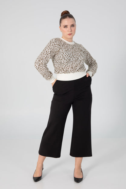 A woman wearing the A25 Pants by Slak in Black, wide legged cropped pants with an elastic waist in Ponte Di Roma fabric. She is wearing them with  the Wanda Sweater by Slak in Leopard and Cream and standing in front of a grey background. 