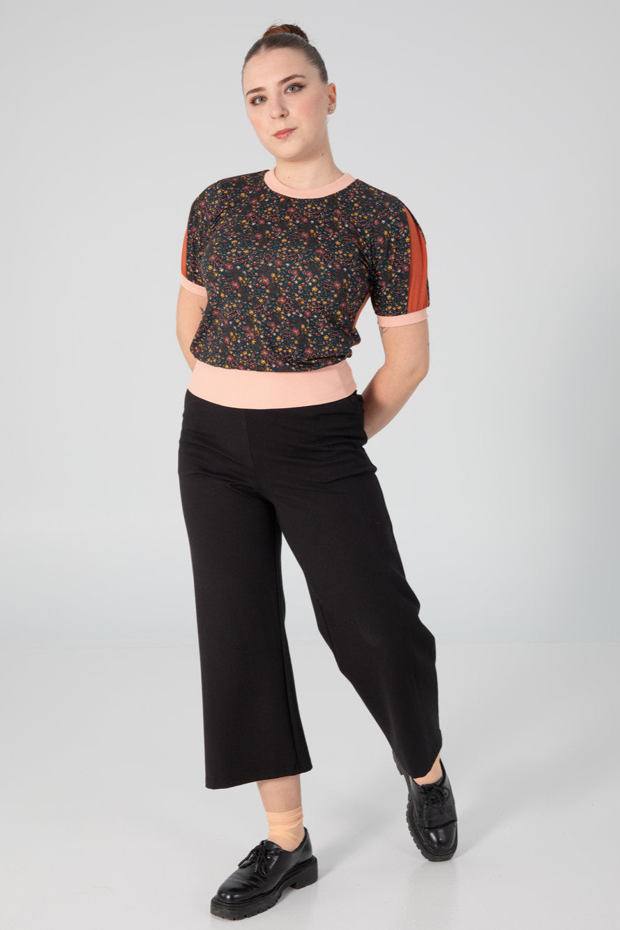 A woman wearing the A25 Pants by Slak in Black, wide legged cropped pants with an elastic waist in Ponte Di Roma fabric. She is wearing them with a patterned top and standing in front of a grey background. 