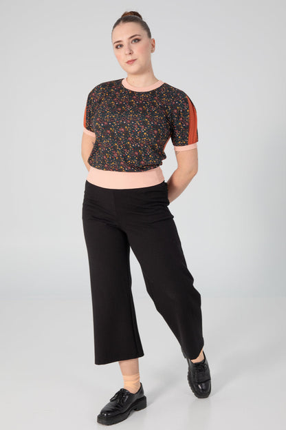 A woman wearing the A25 Pants by Slak in Black, wide legged cropped pants with an elastic waist in Ponte Di Roma fabric. She is wearing them with a patterned top and standing in front of a grey background. 