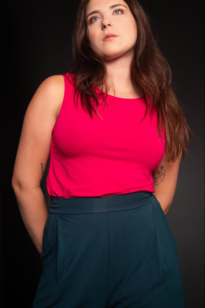 A woman wearing the Neon Camisole by Slak in Fuchsia, a high necked, wide strapped tank. She is wearing it with green pants against a metallic background. 