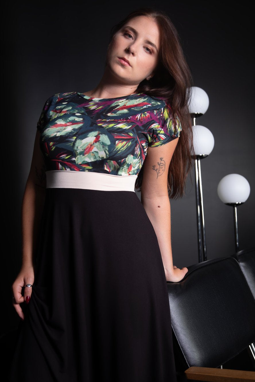 A woman wearing the Capucine Dress by Slak in Black, a fit and flare dress with a leaf print bodice, cream waistband, and black A-line skirt that falls to the knees. It has a round neck in front and a scooped neck at the back. She is standing by a black chair in a dark room. 
