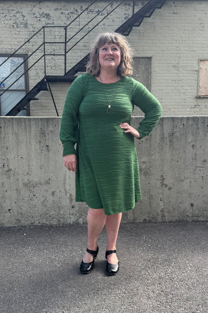 A woman wearing the Long Sleeved Casual Dress FW by SI Design in Green, with a round neck, long cuffed sleeves, and a loose A-line shape. she is standing in front of a building with a staircase.