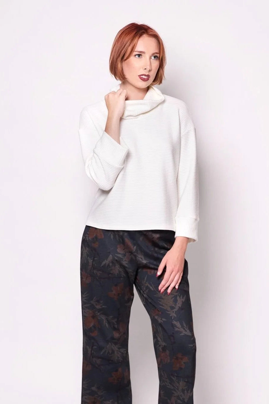 A woman wearing the Sidney Top by Tangente in Ivory, a slightly cropped, boxy top in ribbed fabric with long cuffed sleeves and a large cowl neck. She is wearing it with floral pants and standing in front of a white wall. 