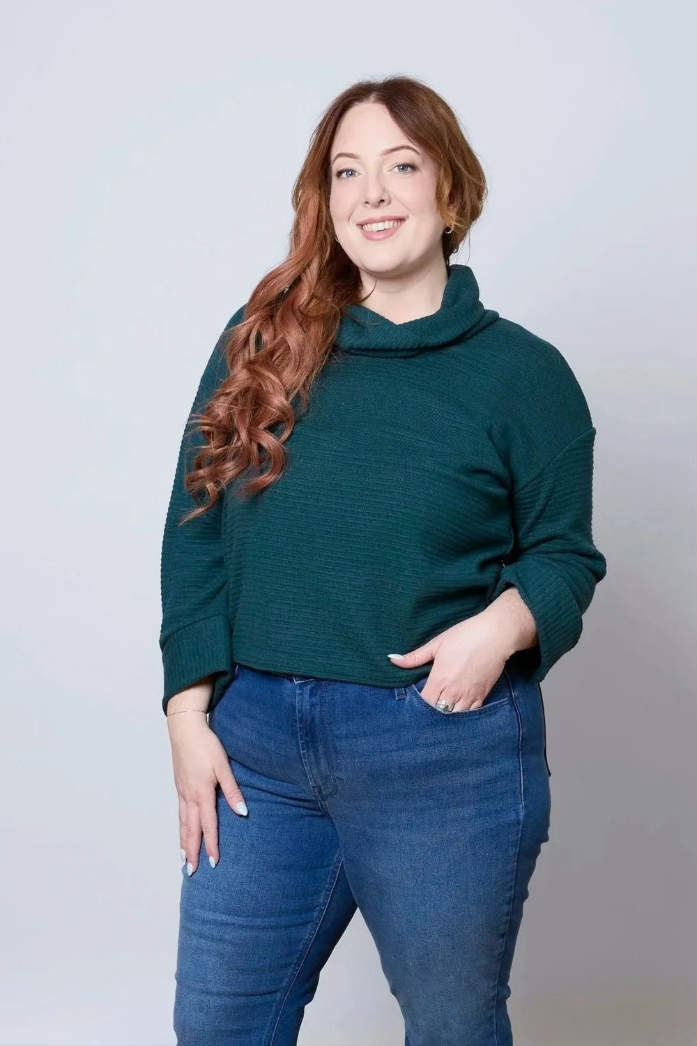 A woman wearing the Sidney Top by Tangente in Pine, a slightly cropped, boxy top in ribbed fabric with long cuffed sleeves and a large cowl neck. She is wearing it with jeans and standing in front of a white wall. 