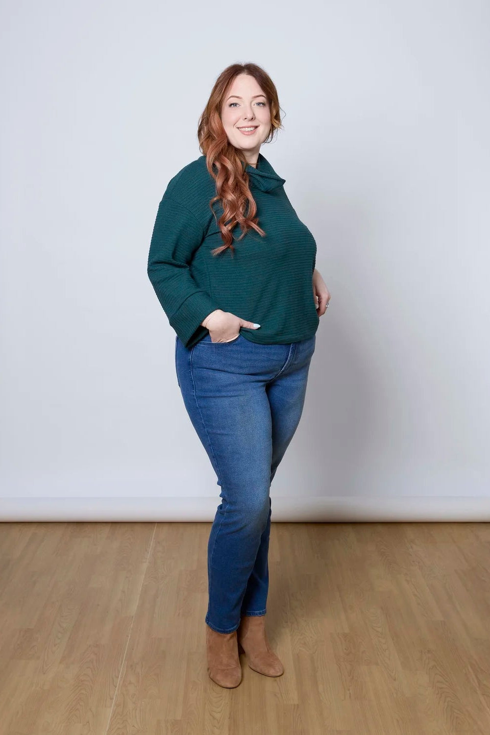 A woman wearing the Sidney Top by Tangente in Pine, a slightly cropped, boxy top in ribbed fabric with long cuffed sleeves and a large cowl neck. She is wearing it with jeans and standing in front of a white wall. 