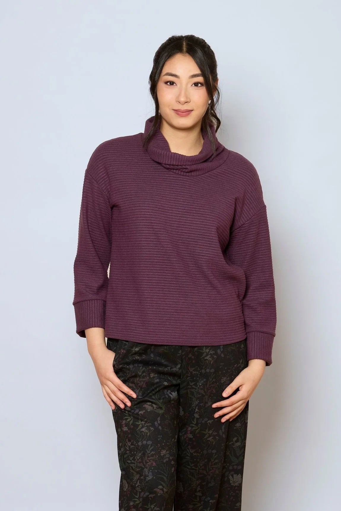 A woman wearing the Sidney Top by Tangente in Purple, a slightly cropped, boxy top in ribbed fabric with long cuffed sleeves and a large cowl neck. She is wearing it with floral pants and standing in front of a white wall. 
