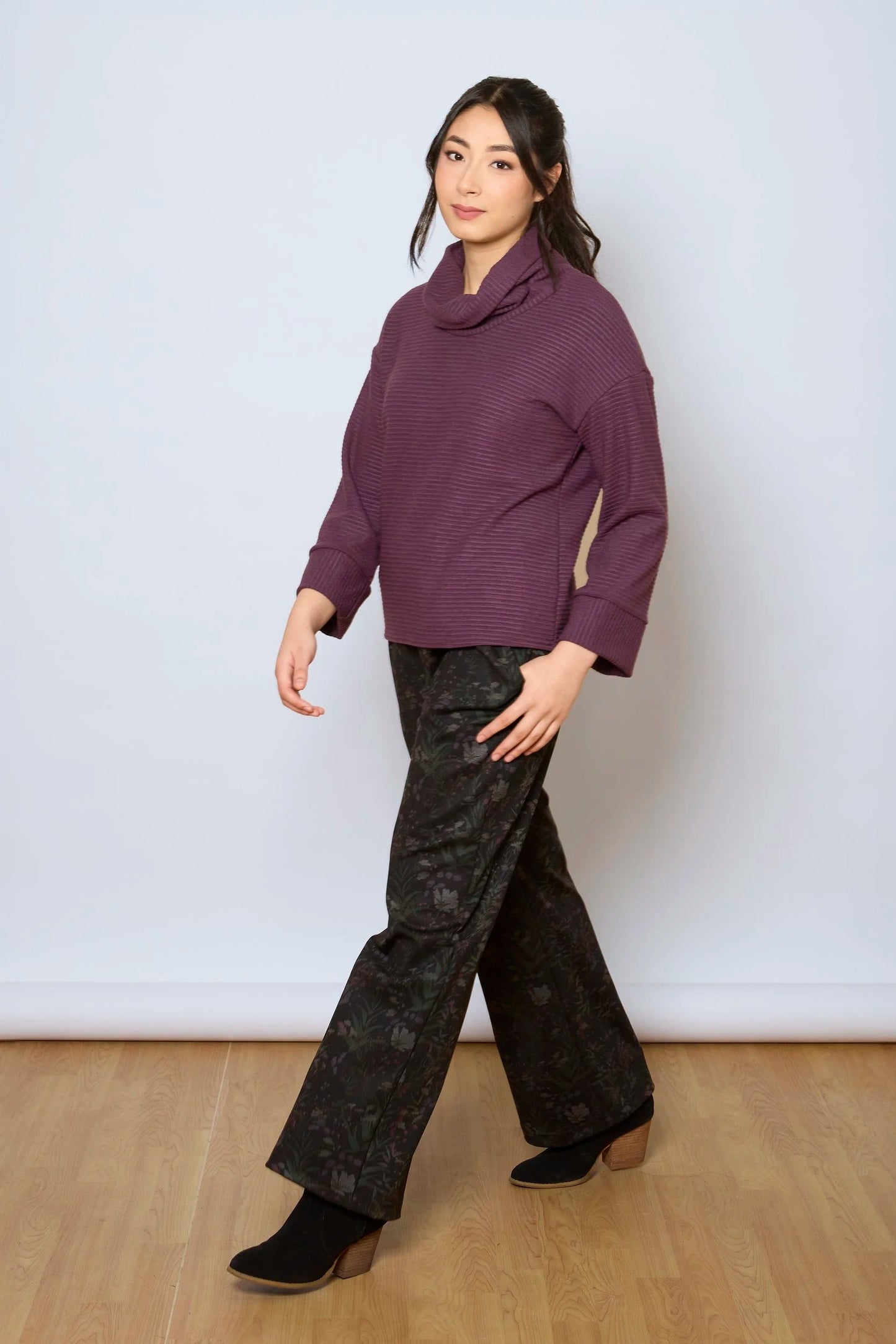 A woman wearing the Sidney Top by Tangente in Purple, a slightly cropped, boxy top in ribbed fabric with long cuffed sleeves and a large cowl neck. She is wearing it with floral pants and standing in front of a white wall. 