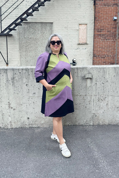 A woman wearing the Lorraine Dress by Solomia in Avocado/Navy/Purple, an A-line colour-blocked dress in stretch denim with 3/4 sleeves and a knee-length hem. She is standing outside in front of a cement wall. 