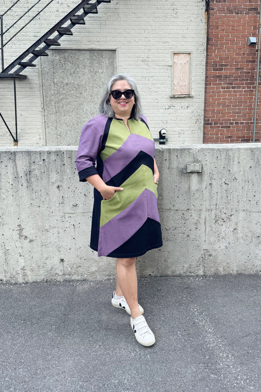 A woman wearing the Lorraine Dress by Solomia in Avocado/Navy/Purple, an A-line colour-blocked dress in stretch denim with 3/4 sleeves and a knee-length hem. She is standing outside in front of a cement wall. 