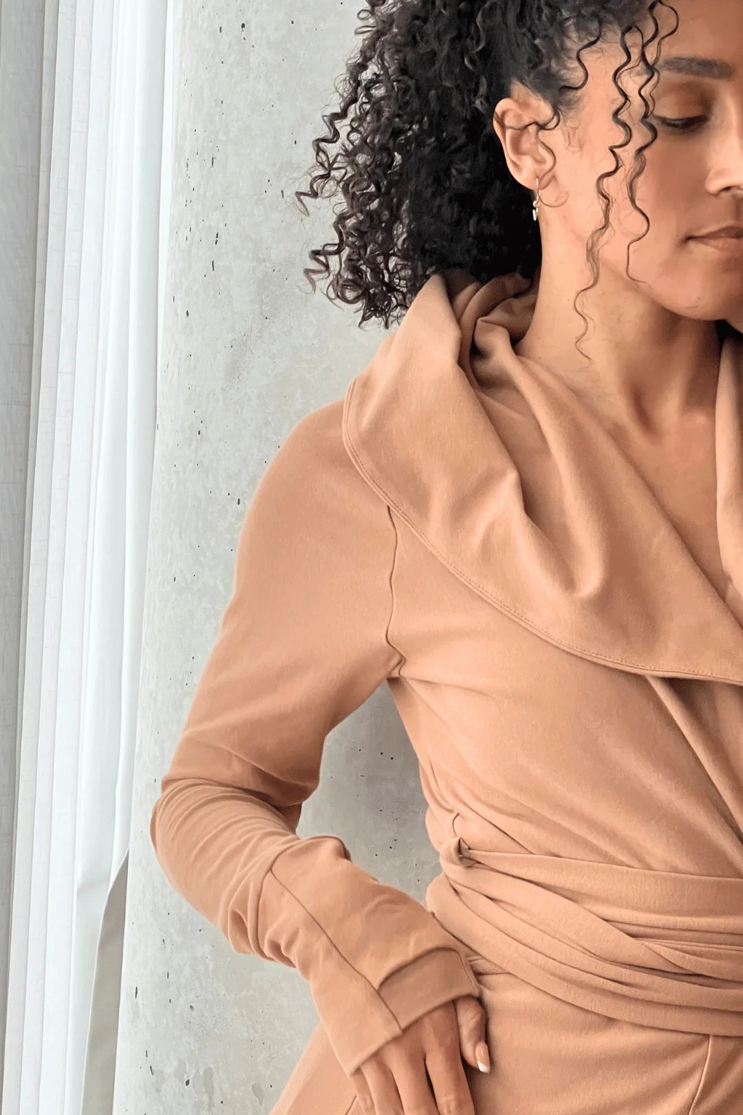 A woman wearing the Signature Cotton Hooded Wrap by Stacey Martin in Caramel, a hooded, corset waist sweatshirt with a tie belt. She is standing against a cement wall. 