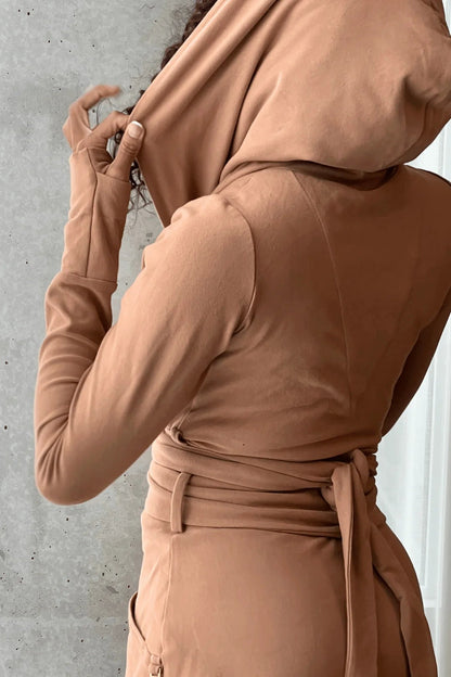 Back view of a woman wearing the Signature Cotton Hooded Wrap by Stacey Martin in Caramel, a hooded, corset waist sweatshirt with a tie belt. She is standing against a cement wall. 