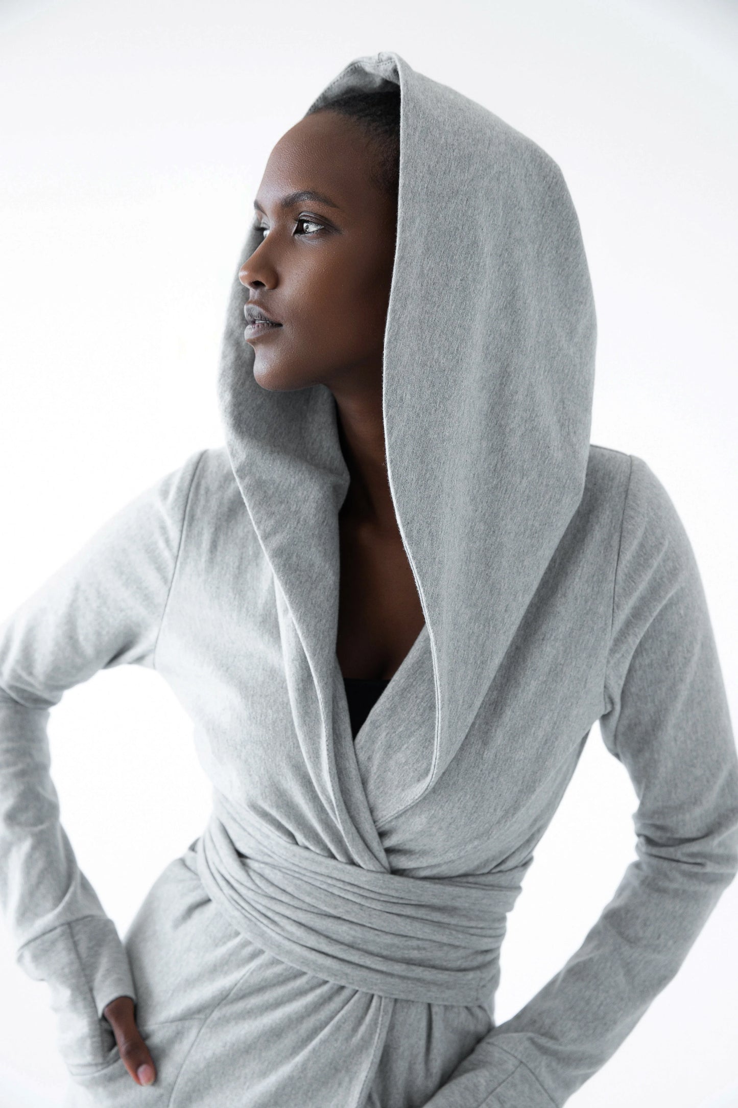 A woman wearing the Signature Cotton Hooded Wrap by Stacey Martin in Stone a hooded, corset waist sweatshirt with a tie belt. She is standing against a white background. 