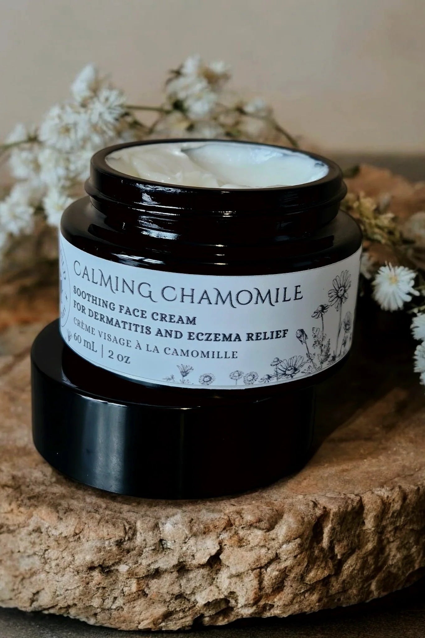 A jar of 'Calming chamomile' face cream by Stay Wild Apothecary is shown on a wooden board with white flowers