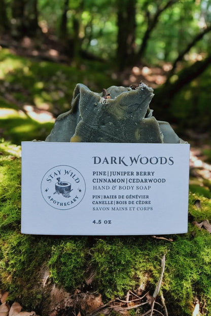 A bar of 'Dark Woods' Soap by Stay Wild Apothecary is shown on moss in a forest setting. 