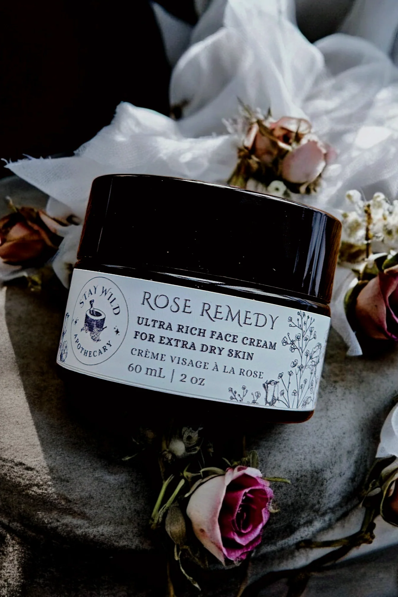 A jar of 'Rose Remedy' ultra rich face cream by Stay Wild Apothecary is shown with flowers on a stone surface