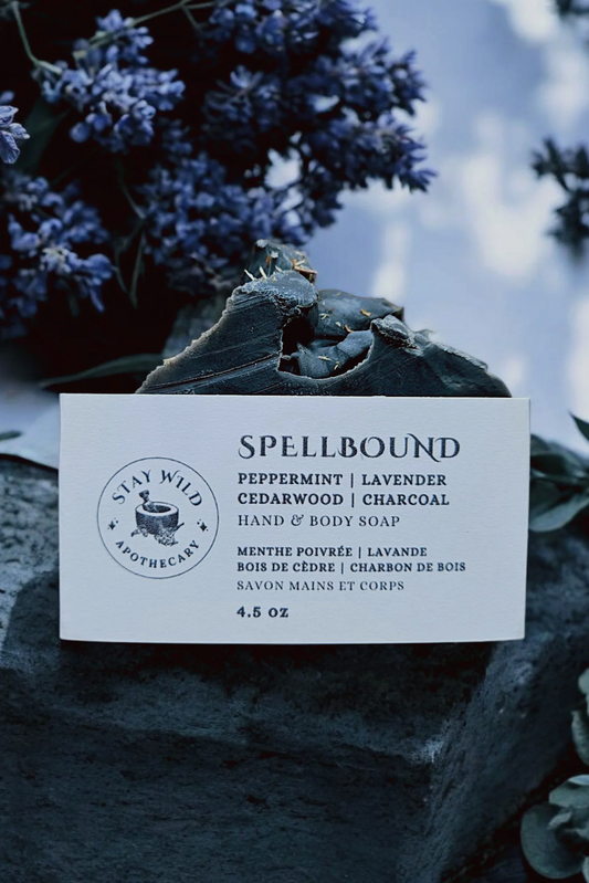 A bar of 'Spellbound' soap by Stay Wild Apothecary is shown on a stone with flowers surrounding it. 