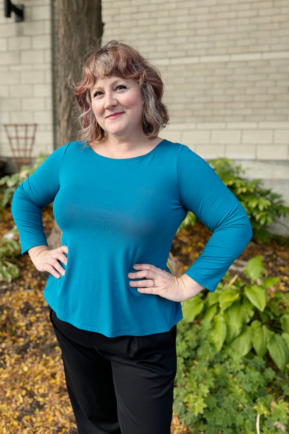 A woman wearing the Studio D Fela top in Teal, featuring 3/4 sleeves and a split hem. She is wearing the Wilfred Pants by Melow in Black and standing in front of a garden. 