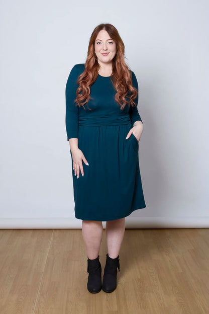 A woman wearing the Cascade Dress by Tangente in Peacock, with a round neck, 3/4 sleeves, gathered defining waistband, and a gathered knee-length skirt. She is standing against a white wall.