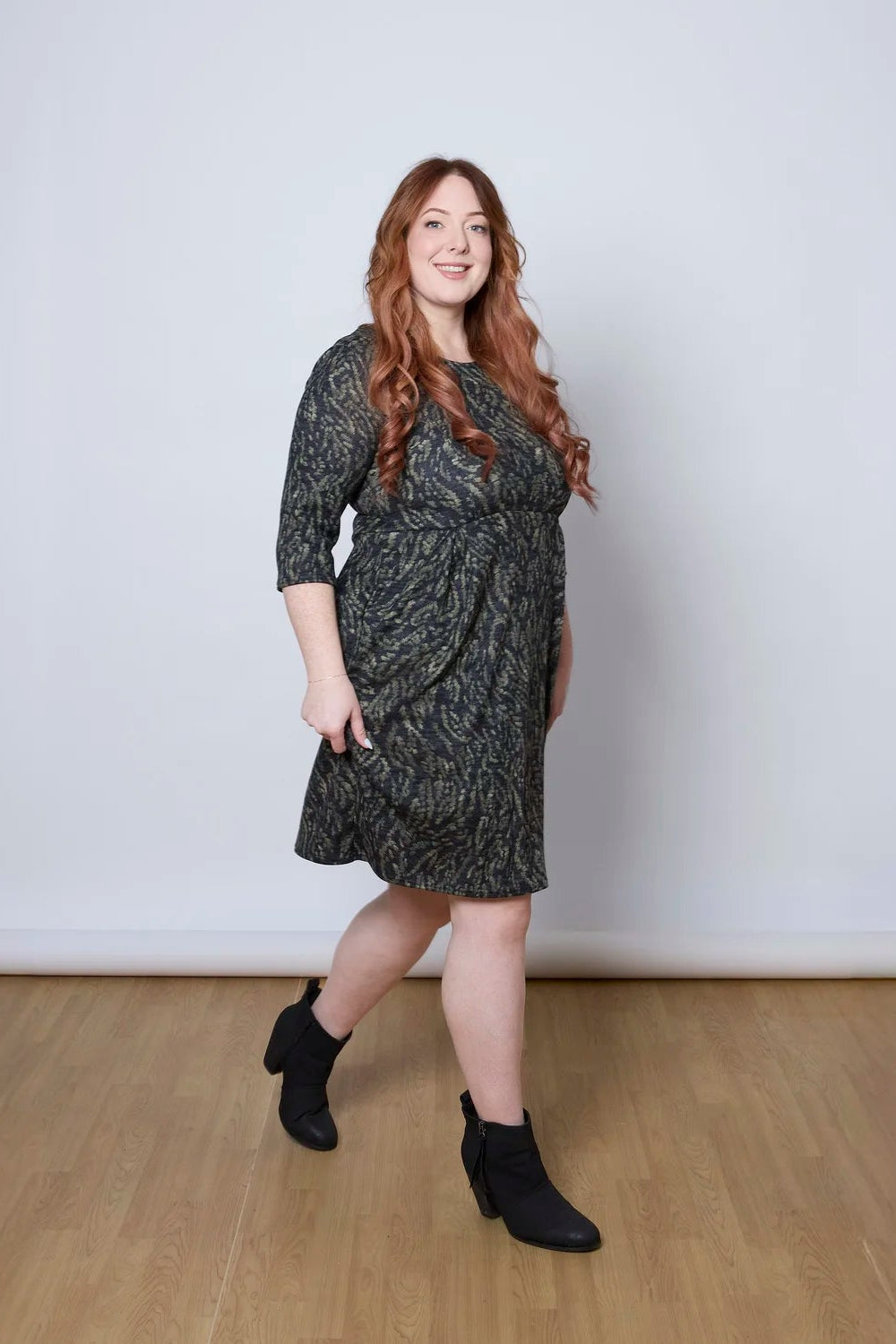 A woman wearing the Meadow Dress by Tangente in Green Print, with a round neck, 3/4 sleeves, princess seams, and a pleated knee-length skirt. She is standing against a white wall. 