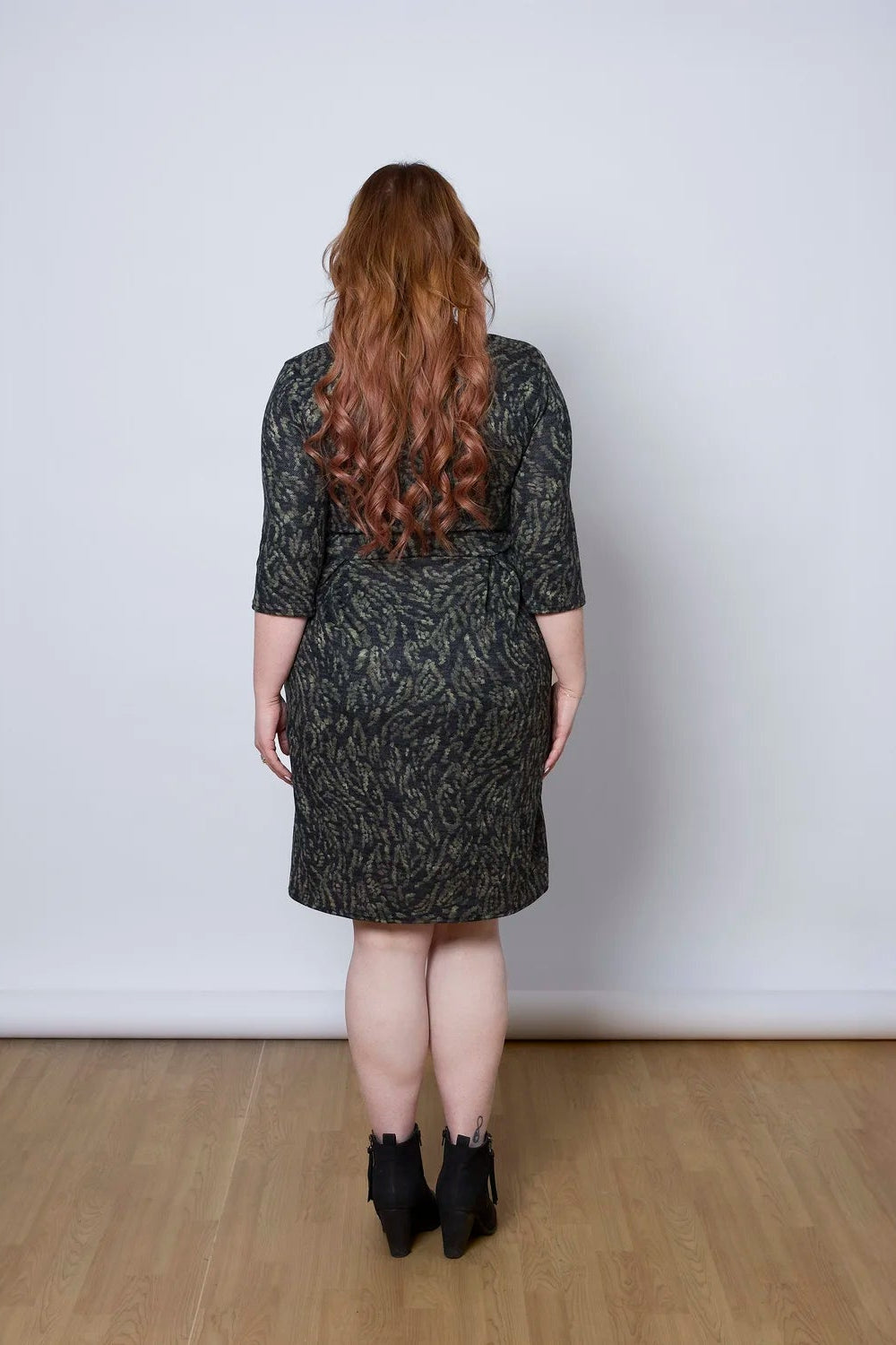 Back view of a woman wearing the Meadow Dress by Tangente in Green Print, with a round neck, 3/4 sleeves, princess seams, and a pleated knee-length skirt. She is standing against a white wall. 