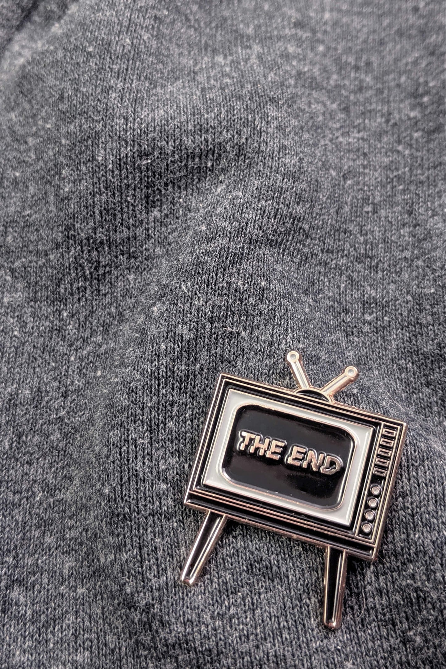  Badaboöm Studio's retro television-shaped enamel pin with 'THE END' lettering on the screen, set against a grey sweatshirt.