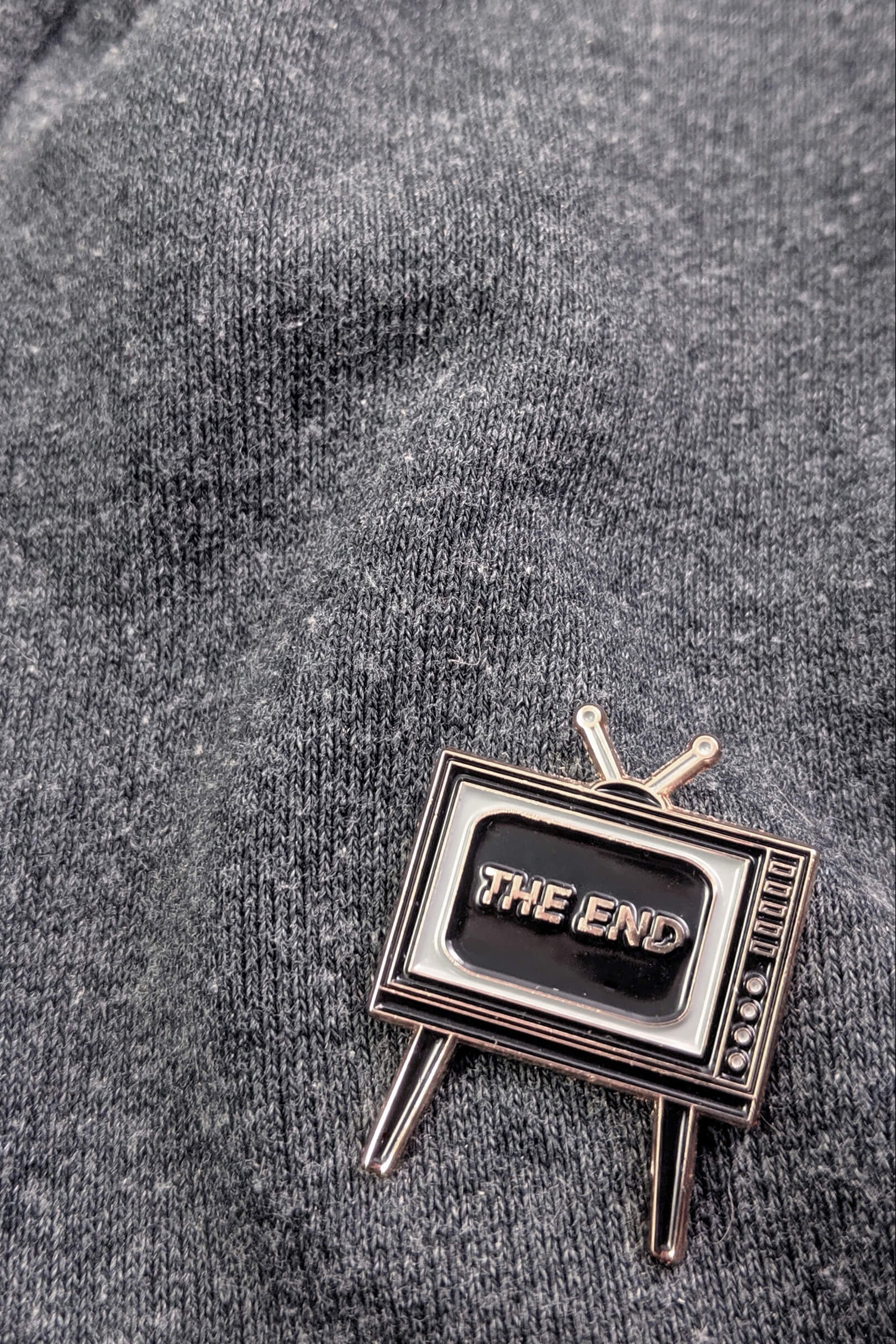  Badaboöm Studio's retro television-shaped enamel pin with 'THE END' lettering on the screen, set against a grey sweatshirt.