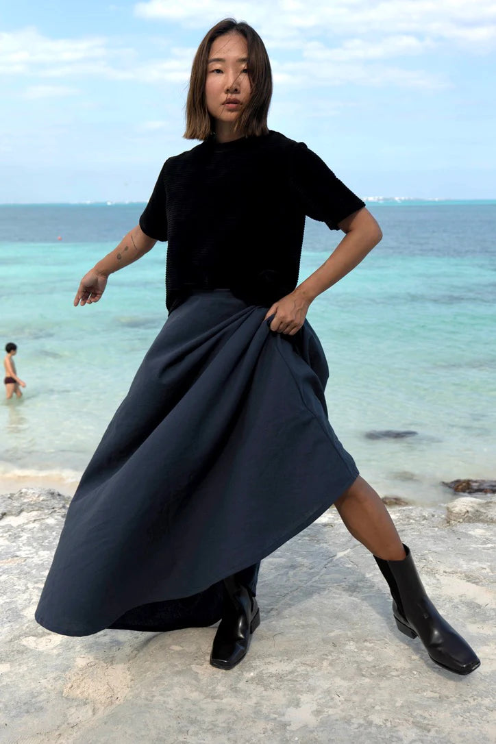 A woman wearing the Tofino T-Shirt in Pepper by MAS, a short sleeved top in ribbed velvet. She is wearing it with a blue skirt and standing on a beach. 