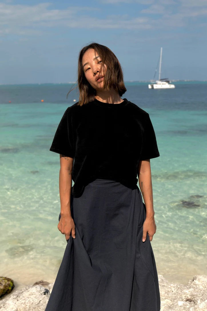 A woman wearing the Tofino T-Shirt by MAS in Pepper, a short sleeved top in ribbed velvet. She is wearing it with a blue skirt and standing on the beach. 