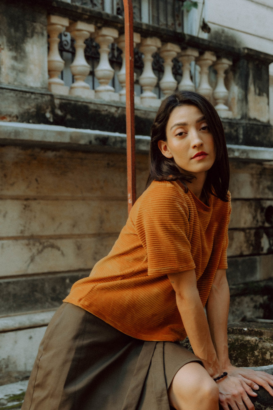A woman wearing the Tofino T-Shirt by MAS in Tumeric, a short sleeved top in ribbed velvet. She is wearing it with a green skirt and sitting in front of a balcony.