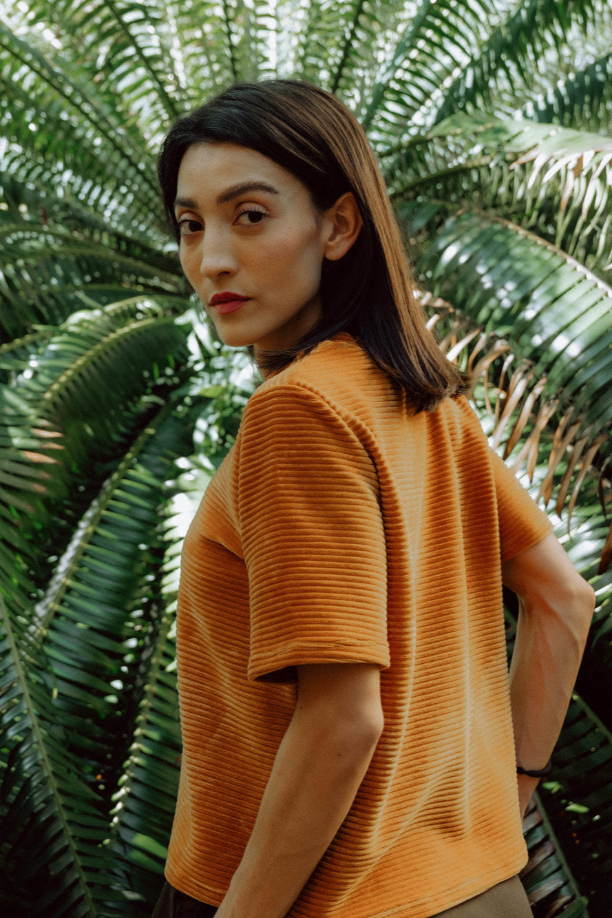 A woman wearing the Tofino T-Shirt in Tumeric by MAS, a short sleeved top in ribbed velvet. She is standing in front of green foliage.