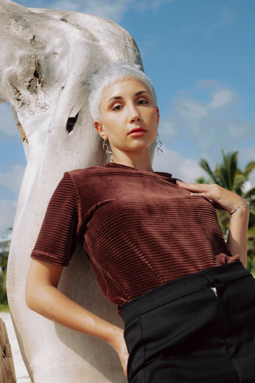 A woman wearing the Tofino T-Shirt by MAS in Cacao, a short sleeved top in ribbed velvet. She is standing on a beach.