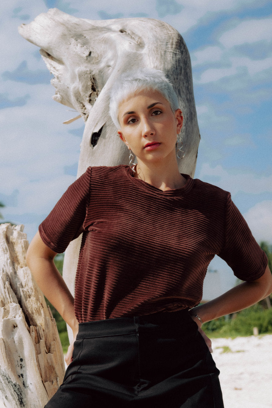 A woman wearing the Tofino T-Shirt by MAS in Cacao, a short sleeved top in ribbed velvet. She is standing on a beach.