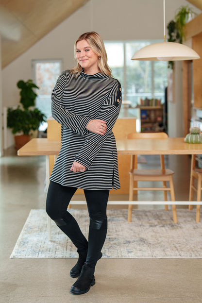 A woman wearing the Aralie Tunic by Rien ne se Perd in Black and White Stripe, with a high neckline, long sleeves, side slits for ease, drop sleeves and a coconut button placket on the left shoulder. She is wearing it with black leggings and standing in a kitchen. 