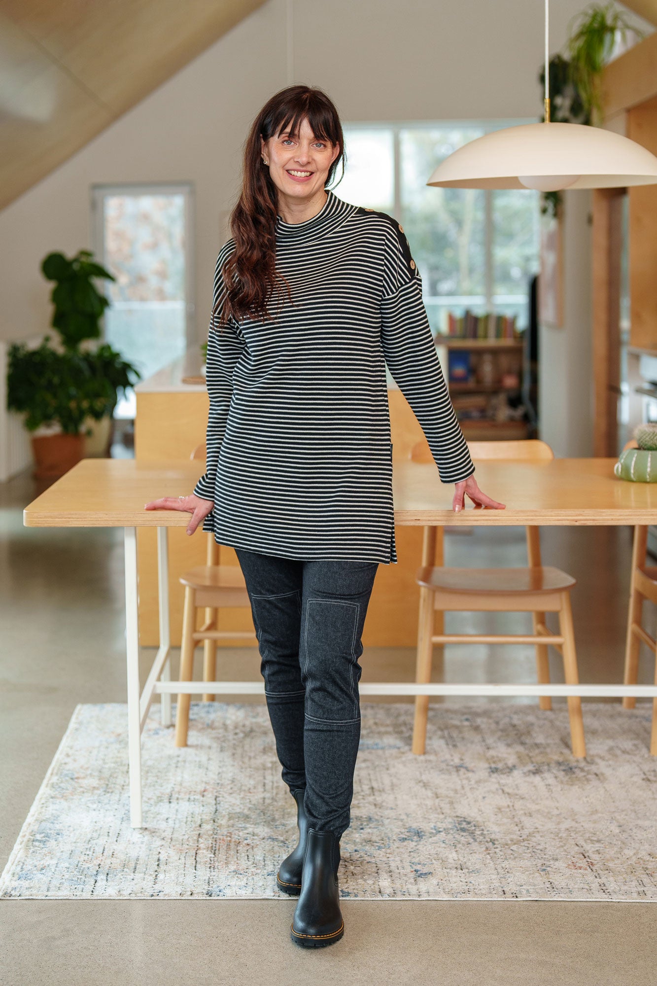 A woman wearing the Cypres Pants by Rien ne se Perd in Grey Denim, stretchy faux denim pants with a pull on waist and tone-on-tone patches on the knees. She is wearing with Aralie Tunic and standing in front of a table.